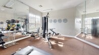 This fitness center offers a thoughtfully designed workout space spread across a single floor. The room is bathed in natural light, courtesy of large multi-pane windows that line the walls and provide views of the outdoor surroundings, creating an open and energizing atmosphere. The interior is finished with neutral, light-toned walls and wall-to-wall carpet flooring, lending the space a clean and comfortable feel.

The cardio area is well-appointed with treadmills and stationary cycling bikes, each equipped with individual display screens for an interactive workout experience. Exercise balls are also on hand for flexibility and core training. On the strength training side, a multi-station cable machine with adjustable benches serves as a centerpiece, complemented by wall-mounted weight plates and a dumbbell rack for a comprehensive resistance workout.

Full-length mirrors line one wall, visually expanding the space and allowing users to monitor their form during exercise. Wall-mounted televisions provide entertainment throughout the room. The entry area includes a small organization station with storage cubbies, sanitation supplies, and waste receptacles, keeping the space tidy and user-friendly. Elegant French doors with glass panels serve as the main entrance, adding a refined architectural touch to this functional and inviting fitness facility.