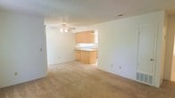 This single-story space features an open-concept design that connects the living area and kitchen in a seamless, functional layout. The living area is finished with wall-to-wall neutral carpeting that lends a warm, understated feel to the space. A ceiling fan with integrated lighting is centrally mounted, providing both air circulation and ambient illumination. The textured ceiling adds subtle visual interest throughout.

The kitchen is positioned openly within the space, defined by light wood cabinetry that extends along the upper and lower sections, offering generous storage. A peninsula-style counter provides a natural divide between the kitchen and living area while also functioning as an additional workspace or casual dining surface. A sink is integrated into the countertop, and the light-toned surfaces throughout contribute to a clean, cohesive aesthetic.

The walls are finished in a crisp, neutral white, complementing the light carpeting and cabinetry. Recessed ceiling vents suggest a central air system. The overall ambiance is bright, clean, and straightforward, making the space a practical and move-in ready environment suited to a variety of living arrangements.
