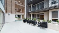 Located at 3401 W Olive Ave in Burbank, California, this contemporary multi-story building showcases a striking dark gray exterior with horizontal metal balcony railings and large-format windows throughout. The architecture incorporates distinctive warm-toned wood-slat screening panels that add visual texture and contrast against the building's modern facade.

The property centers around a series of well-appointed outdoor communal spaces designed for both relaxation and social activity. A ground-level courtyard features polished concrete flooring, sleek black rope-style lounge seating, and raised planters filled with lush tropical greenery, creating a welcoming open-air gathering area. Decorative tilework accents the courtyard floor, adding a refined detail to the space.

An expansive outdoor amenity deck includes a resort-style swimming pool flanked by modern chaise lounges, set against the building's multi-story backdrop. Adjacent to the pool, a generous wood-deck entertaining area provides an outdoor kitchen equipped with multiple built-in grills and a refrigerator, a covered seating lounge, and a mounted outdoor television with fireplace. String lights overhead enhance the ambiance for evening gatherings, while cornhole boards cater to casual recreation. Landscaped planter beds featuring sculptural succulents and drought-tolerant plants border the perimeter, adding a curated natural element throughout.