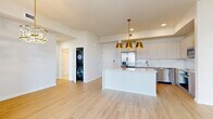 Located along the Pinellas Trail in Saint Petersburg, FL, this contemporary single-floor residence showcases a refined design with a consistent palette of soft whites, warm wood-look flooring, and gold-accented hardware throughout. The open-concept kitchen is a standout feature, centered around a striking marble-veined quartz waterfall island complemented by taupe cabinetry, stainless steel appliances including a French door refrigerator and dishwasher, a marble tile backsplash, and a built-in wine cooler. A trio of gold pendant lights adds warmth and elegance above the island. The living and dining areas are bathed in natural light from expansive floor-to-ceiling sliding glass doors that open to a balcony revealing sweeping skyline views of downtown Saint Petersburg. The bedrooms feature plush carpeting, generous closet space, and large windows that continue the theme of abundant natural light and urban vistas. The bathrooms are finished with light tile flooring, gold-hardware vanities, a glass-enclosed walk-in shower, and a soaking tub with subway tile surround. A stacked in-unit washer and dryer adds everyday convenience. Gold light fixtures and chandelier accents throughout lend a cohesive, upscale aesthetic to the entire space.