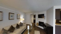 This single-floor residence offers a thoughtfully designed living space that combines comfort with classic style. The living room serves as the heart of the home, anchored by a white-mantled fireplace that adds warmth and a timeless focal point to the room. Rich, dark hardwood flooring runs throughout, providing an elegant contrast to the neutral, light-toned walls and creating a cohesive, inviting atmosphere. A textured ceiling adds subtle architectural interest overhead. The living area is generously proportioned, accommodating a plush sofa ensemble centered around a wooden coffee table, making it well-suited for both relaxing and entertaining. A large flat-screen television is mounted above the fireplace, enhancing the room's functionality. Plantation shutters on the windows offer both privacy and light control, contributing to the space's refined aesthetic. A hallway visible from the main living area leads toward a kitchen featuring dark cabinetry, hinting at additional well-appointed spaces beyond. The overall ambiance is warm and welcoming, with a neutral color palette and classic design elements that create a move-in-ready environment with lasting appeal.