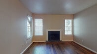 This single-floor residence offers a well-organized layout with consistent hardwood flooring flowing throughout the living spaces, creating a warm and cohesive aesthetic. The living room serves as the focal point of the home, showcasing a classic wood-burning fireplace framed by a clean white surround and a brick hearth. Two generously sized windows fitted with horizontal blinds flank the fireplace, allowing ample natural light to fill the room and highlight the rich, dark-toned hardwood floors. A separate room features a sliding glass door with vertical blinds, opening to an outdoor area and providing a bright, airy connection to the exterior. The kitchen is equipped with white cabinetry, granite-style countertops, and a stainless steel dishwasher, offering both functionality and a clean, modern look with warm wood flooring underfoot. The hallway, lit by a ceiling fan with a light fixture, connects the living spaces to the bedrooms, one of which features neutral tones and the same hardwood flooring. The bathroom includes a tiled shower-tub combination, white cabinetry with overhead storage, granite-topped vanity, and a mirrored medicine cabinet. Throughout the home, neutral wall tones and white trim create a clean, inviting atmosphere.