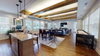 This single-floor community gathering space is thoughtfully designed to blend comfort and functionality in an open, inviting layout. The most striking architectural feature is the ceiling, which showcases a series of bold, exposed wooden beams with warm uplighting set against a crisp white backdrop — creating a rustic-modern aesthetic that anchors the entire room. Rich, wide-plank hardwood flooring runs throughout, adding warmth and continuity across all areas of the space.

The main lounge area is generously proportioned, furnished with comfortable sofas, mid-century style armchairs, and layered area rugs, all arranged to encourage conversation and relaxation. A large wall-mounted flat-screen television serves as a focal point in this zone. Adjacent to the lounge, a dedicated dining and high-top bar seating area provides ample space for casual gatherings or group meetings.

The kitchenette is well-appointed, featuring granite countertops, warm wood cabinetry, a mosaic tile accent wall, pendant lighting, and appliances including a coffee station, microwave, and dishwasher — ideal for entertaining. Multiple large windows allow abundant natural light to pour in throughout the space, offering pleasant outdoor views. A glass-paned French door provides an additional point of entry and further enhances the bright, airy atmosphere of the room.