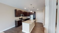 This single-floor space showcases a thoughtfully designed kitchen that serves as the heart of the home. Rich, dark espresso cabinetry lines the walls, providing generous storage while creating a bold contrast against the light granite countertops. A central kitchen island with a built-in sink and matching granite surface offers additional workspace and casual seating potential, illuminated overhead by elegant pendant lights. Stainless steel appliances — including a refrigerator, microwave, and range — complete the modern aesthetic, while a wine cooler adds a refined touch beneath the counter. Recessed lighting throughout ensures the space remains bright and inviting. The flooring features wide-plank dark hardwood, lending warmth and continuity across the kitchen and entry areas. Neutral-toned walls provide a versatile backdrop that complements the cabinetry and finishes. A clean white entry door is visible from the kitchen, connecting the space to the entryway corridor. The open layout and well-considered design details combine to create a functional yet stylish living environment suited for both everyday living and entertaining.