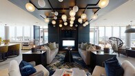 This striking single-floor space showcases a thoughtfully curated interior defined by bold contrasts, designer details, and panoramic city views. The main lounge area commands attention with a dramatic coffered ceiling finished in deep charcoal, adorned with a sculptural installation of globe pendant lights that create a warm, ambient glow. Plush neutral sofas flank a central media wall, while a vibrant abstract area rug anchors the seating arrangement. Blue-and-white striped accent walls lend a graphic energy to the space, complemented by warm wood flooring and cozy banquette nooks with padded leather upholstery.

A separate seating area features built-in wood shelving displaying curated ceramic artworks alongside an integrated flat-screen television, providing a more intimate gathering space. The kitchen is outfitted with a substantial marble-topped island, warm wood cabinetry, and mustard-yellow upholstered bar stools, with floor-to-ceiling windows framing expansive outdoor views.

A bright, glass-walled dining and social area features a large round communal table beneath an artistic sculptural light fixture, opening directly onto a generous outdoor terrace. The terrace offers sweeping city skyline views, hanging egg chairs, and ample space for outdoor lounging, seamlessly blending the interior and exterior environments.