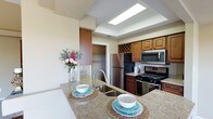 This single-floor residence offers a thoughtfully designed living space centered around a standout kitchen that blends functionality with style. The kitchen is equipped with a full suite of stainless steel appliances, including a refrigerator, gas range, and over-the-range microwave. Rich, warm-toned wood cabinetry lines the walls, complemented by a distinctive built-in wine rack that adds a touch of elegance. Granite countertops extend generously across the workspace, culminating in a spacious kitchen island with a stainless steel sink — ideal for meal preparation and casual dining. A recessed tray ceiling with a skylight panel bathes the kitchen in bright, even light, enhancing the open and airy feel of the space. Adjacent to the kitchen, a walk-in pantry or storage closet provides ample organizational space. The light-toned walls and tile flooring contribute to a clean, neutral palette throughout. Overall, the space presents a cohesive and practical layout that balances everyday convenience with refined design details, making it a welcoming and efficient environment for both cooking and entertaining.
