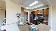 This single-floor residence offers a thoughtfully designed living space centered around a standout kitchen that blends functionality with style. The kitchen is equipped with a full suite of stainless steel appliances, including a refrigerator, gas range, and over-the-range microwave. Rich, warm-toned wood cabinetry lines the walls, complemented by a distinctive built-in wine rack that adds a touch of elegance. Granite countertops extend generously across the workspace, culminating in a spacious kitchen island with a stainless steel sink — ideal for meal preparation and casual dining. A recessed tray ceiling with a skylight panel bathes the kitchen in bright, even light, enhancing the open and airy feel of the space. Adjacent to the kitchen, a walk-in pantry or storage closet provides ample organizational space. The light-toned walls and tile flooring contribute to a clean, neutral palette throughout. Overall, the space presents a cohesive and practical layout that balances everyday convenience with refined design details, making it a welcoming and efficient environment for both cooking and entertaining.