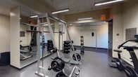 This fitness center offers a comprehensive workout environment thoughtfully organized into distinct training zones. The strength training area is well-appointed with a squat/power rack, a barbell and weight plate setup, a full rack of dumbbells, and a medicine ball tower — all positioned in front of floor-to-ceiling mirrors that enhance the sense of space and allow users to monitor their form. The rubber flooring throughout provides durability and safety underfoot.

The cardio zone features multiple treadmills and upright stationary bikes, with wall-mounted flat-screen televisions positioned directly in front of the cardio machines for entertainment during workouts. Additional cardio and cross-training equipment, including an elliptical trainer and a rowing machine, rounds out this section of the facility.

A dedicated selectorized strength machine station offers multi-function cable and resistance training options, ideal for targeted muscle group exercises. Overhead, linear LED lighting fixtures provide bright, even illumination across the entire space, contributing to a clean and functional atmosphere. The neutral-toned walls and consistent flooring material create a cohesive, professional aesthetic throughout. The facility is well-maintained and designed to support a wide range of fitness goals, from cardiovascular conditioning to strength and functional training.