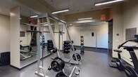 This fitness center offers a comprehensive workout environment thoughtfully organized into distinct training zones. The strength training area is well-appointed with a squat/power rack, a barbell and weight plate setup, a full rack of dumbbells, and a medicine ball tower — all positioned in front of floor-to-ceiling mirrors that enhance the sense of space and allow users to monitor their form. The rubber flooring throughout provides durability and safety underfoot.

The cardio zone features multiple treadmills and upright stationary bikes, with wall-mounted flat-screen televisions positioned directly in front of the cardio machines for entertainment during workouts. Additional cardio and cross-training equipment, including an elliptical trainer and a rowing machine, rounds out this section of the facility.

A dedicated selectorized strength machine station offers multi-function cable and resistance training options, ideal for targeted muscle group exercises. Overhead, linear LED lighting fixtures provide bright, even illumination across the entire space, contributing to a clean and functional atmosphere. The neutral-toned walls and consistent flooring material create a cohesive, professional aesthetic throughout. The facility is well-maintained and designed to support a wide range of fitness goals, from cardiovascular conditioning to strength and functional training.