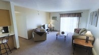 This single-floor space offers a comfortable and functional layout centered around a generously sized living room. The living room features wall-to-wall carpeting that adds warmth underfoot, along with light-toned walls that create a bright, neutral backdrop. A large window dressed with decorative valance curtains allows an abundance of natural light to fill the room, enhancing its open and airy feel. A wall-mounted air conditioning unit ensures year-round comfort. The living area provides ample space for multiple seating arrangements, including plush armchairs and a sofa, centered around a classic coffee table with wrought-iron legs. A dedicated corner accommodates an entertainment setup, complete with a media console. A small side table with a lamp adds a warm, ambient lighting option for evenings. The overall ambiance is modest and practical, with a lived-in charm that makes the space feel approachable and inviting. Clean lines, simple décor, and a straightforward layout make this space easy to personalize and well-suited for everyday living.