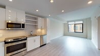 This single-floor unit showcases a clean, modern design with a thoughtfully arranged open layout. The kitchen is a standout feature, equipped with high-gloss white cabinetry, granite countertops, a neutral tile backsplash, and a full suite of stainless steel appliances including a refrigerator, range, built-in microwave, and dishwasher. Open shelving adds a functional, contemporary touch alongside ample closed cabinet storage. The kitchen flows seamlessly into the living area, which is finished with light hardwood-style flooring that runs throughout the main living spaces, contributing to an airy and cohesive feel. Large black-framed windows in the living and adjacent room allow generous natural light and offer views of the surrounding urban streetscape. A separate bedroom features neutral carpet flooring, a large window, and clean white walls, providing a quiet retreat. The bathroom is well-appointed with wood-look tile flooring, a full soaking tub with white subway tile surround accented by a mosaic tile border, a granite-topped vanity, and recessed lighting. Recessed lighting throughout the unit, white walls, and crisp trim work reinforce the overall contemporary and well-maintained character of the space.