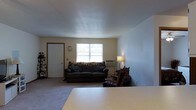 This single-floor space presents a straightforward and functional layout centered around a modest living area. The room is finished with wall-to-wall carpeting in a neutral tone, contributing to a comfortable and understated ambiance throughout. The walls are painted in a light, muted hue that helps maintain a bright and open feel. A centrally positioned window along the far wall allows natural light to filter into the space, illuminating the room and providing an outdoor view. Beneath the window, a window-mounted air conditioning unit provides climate control. The living area is furnished with a dark, cushioned sofa accented with patterned throw pillows, paired with a wooden rocking chair positioned to one side, adding a classic touch to the room. A traditional floor lamp beside the seating area offers warm supplemental lighting. An interior door with wood trim framing is visible along one wall, offering access to adjacent areas of the home. A wall clock is mounted near the door, adding a practical decorative element. A white table surface is partially visible in the foreground, suggesting additional functional space within the room. Overall, the space offers a simple and livable environment with essential amenities.