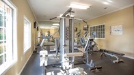 The fitness center at Holly Point, located at 2540 Holly Point Blvd in Chesapeake, Virginia, is a dedicated single-floor exercise facility designed to support a well-rounded workout routine. The space is accessed via a tiled hallway that leads directly into the gym area, providing a clean and organized transition into the facility.

The main workout area features durable rubber flooring throughout, offering both safety and comfort during exercise. The room is finished with warm beige walls, crisp white crown molding, and bright overhead fluorescent lighting, creating a well-lit and functional environment. A large wall-mounted mirror runs along one side of the room, visually expanding the space and allowing users to monitor their form during workouts.

The facility is equipped with a comprehensive selection of cardio machines, including treadmills, ellipticals, and additional aerobic equipment arranged in an orderly row. On the strength training side, a multi-station cable machine, free weights, and adjustable benches are available, accommodating a variety of resistance exercises. A dumbbell rack with a full range of weights is positioned along one wall, complemented by a posted Vectra exercise guide for user reference.

Natural light filters in through multiple windows, enhancing the open feel of the space. A glass-paneled exterior door provides an additional access point. The facility is clean, well-maintained, and thoughtfully organized to serve the fitness needs of the Holly Point community.