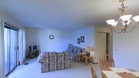 This single-floor space offers a practical and comfortable layout designed for everyday living. The living area serves as the heart of the home, featuring plush wall-to-wall carpeting that adds warmth underfoot. A sectional sofa with patterned upholstery provides generous seating, complemented by a side table with a lamp that adds a soft, ambient glow to the room. A wall-mounted clock and framed artwork add simple decorative touches to the neutral-toned walls, while a television set on a low media stand anchors one corner of the room. The textured ceiling adds a subtle architectural detail overhead. Adjacent to the living area, a dining space is visible, outfitted with wooden chairs that offer a natural contrast to the carpeted living zone. A shelving unit can be spotted in the background, providing practical storage options. The open flow between the living and dining areas creates a sense of spaciousness, making the layout feel connected and easy to navigate. Overall, the space presents a cozy, no-fuss environment with a neutral color palette that offers a calm and welcoming ambiance throughout.