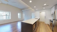 Located at 1220 Huron Rd E in Cleveland, Ohio, this modern single-floor unit offers a thoughtfully designed open-concept layout with contemporary finishes throughout. The living area is bright and spacious, featuring warm hardwood-style flooring, soaring ceilings with exposed beam detailing, and multiple windows that frame urban views of the surrounding cityscape.

The kitchen is a standout feature, showcasing a large center island with a white quartz countertop and built-in microwave drawer, complemented by sleek dark cabinetry. The full kitchen wall features white upper cabinets, a tile backsplash, stainless steel appliances including a range and refrigerator, and a range hood — all illuminated by recessed lighting above.

The bedroom is generously proportioned with vaulted ceiling elements, hardwood-style flooring, and a large window providing natural light and city views. It connects directly to a walk-in closet with built-in shelving.

The bathrooms are finished in a clean, neutral palette with large-format light tile flooring. One bathroom includes a soaking tub with a tiled surround and handheld showerhead, while another features a glass-enclosed shower. Both include floating vanities with white countertops and accessibility grab bars. An in-unit washer and dryer is also included.
