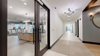 This expansive single-floor amenity space offers a thoughtfully designed environment that blends fitness, socializing, and relaxation across a generous footprint. The facility is anchored by a large, well-appointed exercise room featuring a wide array of equipment, including power racks with weight plates, cardio machines such as treadmills and stair climbers, a punching bag, suspension trainers, and a central wooden island counter — all beneath sleek linear pendant lighting and set on durable, easy-clean flooring. Wide glass-paneled doors with dark metal frames provide clear sightlines into the fitness area from the hallway, creating an open and inviting aesthetic. The hallways themselves are bright and polished, finished with large-format tile floors, dark wood accents, geometric chandelier lighting, and decorative touches like a round gold-framed mirror. A comfortable lounge area with upholstered outdoor-style seating opens to a covered patio with a wood-slat bar counter, bar stools, and a mounted TV — ideal for communal gatherings. A private meeting room features a central conference table, modern pendant lighting, textured wall coverings, and a large window with natural light. The exterior entry corridor showcases stone cladding and landscaped surroundings, reflecting the community's polished, contemporary character throughout.