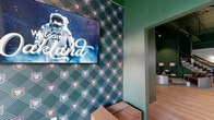 Located at 2801 Locust St in St. Louis, Missouri, this single-floor commercial space makes a strong first impression with its dramatic interior design. Deep forest green walls dominate throughout, creating a rich, immersive atmosphere that is both sophisticated and energetic. Light hardwood flooring stretches across the open layout, providing a warm contrast to the bold wall color and the exposed industrial ceiling above, which features visible structural beams and ductwork painted to match the green palette.

The main area is generously proportioned, offering an open, flexible floor plan well-suited for a variety of uses. A grand staircase with a dark wood handrail and wrought iron balusters serves as a striking architectural centerpiece, leading to upper levels. An elevator is also accessible, adding convenience.

The space is furnished with distinctive, eclectic seating — including curved metallic sofas and sculptural accent chairs — lending a contemporary, lounge-like character. Multiple flat-screen displays are mounted along one wall, ideal for digital signage or entertainment. Elegant wall sconces illuminate a clean white accent wall, adding a refined touch. Natural light filters in through glass-paned entry doors at one end, while recessed and track lighting illuminate the interior throughout. The overall ambiance is bold, modern, and highly curated.