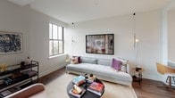 Located at 538-546 Broad St in the heart of Newark, NJ, this studio and den unit delivers a thoughtfully designed single-floor layout that maximizes functionality and style. The open-concept living area features rich hardwood flooring, crisp white walls, and large grid-pane windows that frame elevated views of the Newark cityscape, filling the space with natural light. The living zone offers generous room for both lounging and entertaining, with a clean, modern aesthetic throughout.

The kitchen is outfitted with light wood cabinetry, sleek white quartz countertops, stainless steel appliances including an electric range with over-the-range microwave, and a subtle tile backsplash — combining practicality with contemporary design.

A distinctive feature of this unit is the textured concrete-finish accent walls found in both the den and the sleeping area, lending an urban, loft-inspired character. The den provides a versatile separate space, ideal as a home office or additional sleeping area, and is complemented by its own cozy atmosphere.

The bathroom is finished with herringbone-patterned tile flooring, a modern floating vanity with warm wood cabinetry, and a clean, all-white surround, creating a polished and serene environment. The overall ambiance of the unit balances industrial-chic design elements with modern residential comfort.