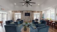 Located at 3050 Plantation Garden Way in Fayetteville, NC, this single-floor community clubhouse delivers a polished, coastal-inspired environment designed for both relaxation and productivity. The centerpiece of the space is a generously sized lounge area boasting soaring vaulted ceilings, hardwood-style flooring, and a striking reclaimed wood accent wall that anchors a wall-mounted flat-screen TV. Plush navy velvet seating is arranged around glass-topped coffee tables atop vivid teal-toned area rugs, creating an inviting and visually dynamic gathering space. Large French doors and windows flood the room with natural light. An adjacent dining area features a substantial solid wood farmhouse table with matching chairs, accented by bold abstract artwork and crown molding. The well-appointed kitchenette offers white quartz countertops, dark cabinetry, stainless steel appliances, and a vibrant teal glazed brick backsplash. The business center provides dedicated workstations with computers and printing equipment, complemented by calming blue-gray walls and contemporary artwork. A quieter alcove offers a single seating chair with decorative wall-mounted glass orbs, ideal for focused tasks. Throughout, hardwood-style flooring, curated artwork, and a cohesive teal-and-blue palette unify the space with a refined, welcoming ambiance.