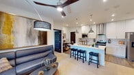 This well-appointed single-floor residence showcases a thoughtfully designed open-concept layout that seamlessly connects the living, dining, and kitchen areas. The living room features wood-look flooring, a striking large-blade ceiling fan, and natural light filtering through slatted blinds, creating a warm and inviting atmosphere complemented by bold artwork and a navy leather sectional sofa.

The kitchen is a standout feature, boasting crisp white shaker cabinetry, a subway tile backsplash, stainless steel appliances including a range with a wall-mounted hood, and an expansive waterfall-edge quartz island with seating for multiple guests. Industrial-style pendant lights hang above the island, adding a modern edge to the space.

The primary bedroom offers a comfortable retreat with carpet flooring, a ceiling fan, natural light from a window, and direct access to a walk-in closet. The en-suite bathroom features large-format tile flooring, a wide dual vanity with white cabinetry, a well-lit mirror, and a glass-enclosed shower. A second bedroom provides ample space with its own window, ceiling fan, and built-in shelving. A second full bathroom echoes the same clean, contemporary aesthetic with neutral tile and a streamlined vanity.
