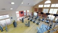 This two-floor fitness center is designed to support a comprehensive range of workout activities in a bright, welcoming environment. The main entrance greets visitors with soaring vaulted ceilings, warm terracotta-toned accent walls, and decorative artwork, setting an inviting tone throughout the facility.

The primary workout floor is generously proportioned, featuring soaring ceilings with clerestory windows that flood the space with natural light. It houses an extensive selection of cardio equipment — including treadmills, ellipticals, stationary bikes, and stair climbers — alongside a full array of strength and resistance machines. Multiple wall-mounted televisions are positioned throughout for entertainment during workouts. Patterned carpeting adds a distinctive design element to the main floor.

A dedicated spin studio offers a focused cycling experience, featuring multiple spin bikes, a wall-mounted television, and large windows with views to the outdoors. Its terracotta accent wall echoes the design language seen throughout the facility.

A separate multi-purpose studio on the upper level features rich hardwood flooring, a cathedral ceiling with a bold accent wall, and ample natural light from a row of windows. This versatile space is suitable for group fitness classes or personal training sessions. A staircase connects the two floors, completing this well-rounded fitness facility.