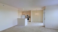 This single-floor space showcases an open-concept layout that seamlessly connects the kitchen, dining, and living areas into one cohesive environment. The kitchen is equipped with white cabinetry, white appliances including a refrigerator, electric range, and microwave, and features a peninsula that provides additional counter space while defining the boundary between the kitchen and the adjacent living area. Recessed lighting illuminates the kitchen zone, offering functional brightness throughout.

The dining area is distinguished by a classic chandelier that adds a traditional decorative touch, hanging elegantly above what would serve as a dining space. The living area is finished with neutral, light-colored carpeting that extends throughout the open space, contributing to a warm and cohesive aesthetic. Soft, neutral wall tones enhance the airy, understated character of the interior.

Natural light enters through what appears to be a sliding glass door or large window on the far wall, brightening the living space and providing a sense of openness. The overall ambiance is clean, neutral, and functional, making it a versatile and welcoming space.