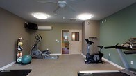 This single-floor fitness room is thoughtfully designed to provide a functional and comfortable workout environment. The space is outfitted with a selection of cardio equipment, including an elliptical trainer, a stationary upright bike, and a treadmill, offering a variety of options for cardiovascular exercise. A resistance band station mounted on the wall adds versatility for strength and flexibility training. The room is finished with neutral-toned walls in shades of tan and sage green, creating a calm and focused atmosphere. Overhead, dual flush-mount light fixtures provide bright, even illumination throughout the space, while a ceiling fan promotes air circulation during workouts. A wall-mounted mini-split air conditioning unit ensures climate comfort year-round. A wall-mounted television adds an entertainment option for those exercising. The flooring is a low-pile carpet suited for gym use. A windowed interior door leads to an adjacent area, contributing a sense of openness. The layout is efficiently arranged to maximize usable floor space, making the room practical for individual fitness routines. Overall, the space is clean, well-lit, and purpose-built for health and wellness.