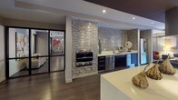 Located at 70 Upper Rock Circle in Rockville, Maryland, this contemporary single-floor amenity space offers a sophisticated and cohesive design throughout. The open-concept layout seamlessly connects a fully equipped kitchen with a spacious lounge and entertainment area, creating an inviting environment for gathering and relaxation.

The kitchen is a standout feature, showcasing a striking textured stone tile accent wall that frames stainless steel appliances, including a built-in wall oven. Dark cabinetry is paired with crisp white countertops, a undermount sink, and a large center island that anchors the space. The tile backsplash continues along the counter wall, adding visual depth and texture.

The lounge area is warm and well-appointed, with light hardwood flooring, recessed lighting, and artfully designed wall niches that display sculptural pieces. Vibrant artwork adds bold color against neutral walls, while a curved accent ceiling detail adds architectural interest. A dedicated media area features a wall-mounted flat-screen television, a low-profile media console, and flexible seating arrangements including ottomans, poufs, and lounge chairs.

Floor-to-ceiling glass doors open directly to an outdoor patio, flooding the interior with natural light and providing seamless indoor-outdoor connectivity. The patio, visible through the expansive glazing, features seating and colorful umbrella-shaded tables, extending the functional space outdoors.