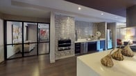 Located at 70 Upper Rock Circle in Rockville, Maryland, this contemporary single-floor amenity space offers a sophisticated and cohesive design throughout. The open-concept layout seamlessly connects a fully equipped kitchen with a spacious lounge and entertainment area, creating an inviting environment for gathering and relaxation.

The kitchen is a standout feature, showcasing a striking textured stone tile accent wall that frames stainless steel appliances, including a built-in wall oven. Dark cabinetry is paired with crisp white countertops, a undermount sink, and a large center island that anchors the space. The tile backsplash continues along the counter wall, adding visual depth and texture.

The lounge area is warm and well-appointed, with light hardwood flooring, recessed lighting, and artfully designed wall niches that display sculptural pieces. Vibrant artwork adds bold color against neutral walls, while a curved accent ceiling detail adds architectural interest. A dedicated media area features a wall-mounted flat-screen television, a low-profile media console, and flexible seating arrangements including ottomans, poufs, and lounge chairs.

Floor-to-ceiling glass doors open directly to an outdoor patio, flooding the interior with natural light and providing seamless indoor-outdoor connectivity. The patio, visible through the expansive glazing, features seating and colorful umbrella-shaded tables, extending the functional space outdoors.