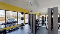 This modern fitness center boasts a thoughtfully designed layout that maximizes functionality and user comfort. The space features high vaulted ceilings with white paint, creating an open and airy atmosphere. Large windows line the walls, flooding the area with natural light and offering views of the surrounding property.

The center's walls are painted a vibrant yellow, adding energy and warmth to the environment. Dark gray flooring provides a durable surface for heavy equipment and high-traffic areas. A variety of exercise equipment is strategically placed throughout the room, including weight machines, free weights, cardio equipment, and stability balls.

The facility is equipped with modern amenities such as ceiling fans for air circulation, wall-mounted televisions for entertainment, and mirrors to assist with proper form during workouts. Multiple entry points, including glass doors, ensure easy access and traffic flow.

Storage solutions for smaller equipment like dumbbells and exercise balls are integrated into the space. The layout appears to be designed to accommodate different workout zones, allowing for simultaneous use by multiple gym-goers. Overall, this fitness center combines functionality with an inviting aesthetic, creating an ideal environment for physical activity and health maintenance.