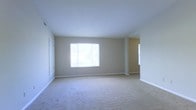 This single-floor space presents a spacious, open living area designed with simplicity and functionality in mind. The room is finished with wall-to-wall beige carpeting that extends throughout, providing a soft, neutral foundation that complements the light gray-beige walls. A large window along one wall allows generous natural light to fill the space, creating a bright and airy atmosphere. White baseboards run consistently along the perimeter, adding a clean, finished look to the interior. The smooth, flat ceiling contributes to an uncluttered aesthetic throughout the room. An open passageway visible toward the rear of the space suggests a connected layout, with an additional area or room accessible beyond the main living space. A secondary window is partially visible near this passageway, further enhancing the flow of natural light. A door is positioned along the left wall, likely providing access to a closet or secondary room. The overall design is understated and versatile, offering a blank canvas well-suited for a variety of furnishing arrangements. The space conveys a functional, move-in ready condition with a clean and well-maintained interior.