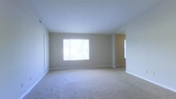 This single-floor space presents a spacious, open living area designed with simplicity and functionality in mind. The room is finished with wall-to-wall beige carpeting that extends throughout, providing a soft, neutral foundation that complements the light gray-beige walls. A large window along one wall allows generous natural light to fill the space, creating a bright and airy atmosphere. White baseboards run consistently along the perimeter, adding a clean, finished look to the interior. The smooth, flat ceiling contributes to an uncluttered aesthetic throughout the room. An open passageway visible toward the rear of the space suggests a connected layout, with an additional area or room accessible beyond the main living space. A secondary window is partially visible near this passageway, further enhancing the flow of natural light. A door is positioned along the left wall, likely providing access to a closet or secondary room. The overall design is understated and versatile, offering a blank canvas well-suited for a variety of furnishing arrangements. The space conveys a functional, move-in ready condition with a clean and well-maintained interior.