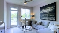 This single-floor residence offers a clean, contemporary aesthetic defined by an abundance of natural light and thoughtful design. The living area serves as the heart of the space, centered around a sleek glass-top coffee table and a plush light-toned sectional sofa, creating an inviting atmosphere perfect for both relaxing and entertaining. A ceiling fan with an integrated light fixture keeps the room comfortable while adding a functional design element overhead. Large windows fitted with horizontal blinds flood the interior with daylight, while a sliding glass door opens to a balcony, framing lush palm trees and a striking urban skyline beyond — bringing the outdoors in with ease. The walls are finished in soft, neutral tones that complement the light-colored flooring, lending the entire space an airy, open feel. A bold large-format artwork depicting a dramatic seascape adds a focal point to the room, infusing the space with visual depth and character. Elegant metallic accent lamps and decorative candle holders contribute refined touches throughout. The overall ambiance is modern, bright, and serene, with a coastal-inspired sensibility that feels both polished and welcoming.