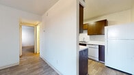This single-floor residence offers a practical and comfortable layout with wood-style flooring carried consistently throughout the home, lending a cohesive and clean aesthetic to every room. The living area is bright and open, featuring a full-light sliding glass door dressed with vertical blinds that invite natural light and provide access to the outdoors. A front entry door with a classic six-panel design anchors the entryway, completing the welcoming feel of the main living space. The kitchen is thoughtfully laid out, featuring warm brown cabinetry both above and below the counters, a double-basin sink, light speckled countertops, and a full suite of appliances including a range, dishwasher, and refrigerator. A half-wall peninsula separates the kitchen from the adjacent dining or living area, creating an open flow between spaces. The bedrooms are generously sized with clean white walls, wood-style flooring, and windows that allow natural light to filter in — one bedroom features a window with an exterior view of surrounding greenery. The bathroom is finished with white tile in the tub/shower surround, wood-style flooring, and straightforward fixtures. A hallway with closet storage connects the rooms, completing this well-organized and functional floor plan.
