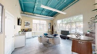 Located at 9001 Kempwood Drive in Houston, TX, this single-floor lounge space makes a striking first impression with its soaring ceilings showcasing boldly painted teal exposed structural beams and ductwork — a design-forward industrial touch that adds character and height to the room. Large windows line multiple walls, flooding the interior with abundant natural light and offering views of the surrounding landscaping and neighborhood.

The living area is thoughtfully arranged with plush, modern lounge chairs in soft gray accented by vibrant teal throw pillows, centered around a geometric patterned ottoman and wire-frame coffee table. A coordinating patterned sofa adds additional seating, while high-top tables with bar stools along the perimeter provide flexible workspace or casual dining options.

Light gray tile flooring runs throughout, contributing to the clean, open aesthetic. A kitchenette area along one wall features white cabinetry, a countertop with sink, a beverage refrigerator, and a coffee station — ideal for entertaining or everyday use. A wall-mounted flat-screen TV completes the space. Warm beige walls paired with contemporary artwork create a welcoming, well-curated ambiance throughout this versatile communal space.