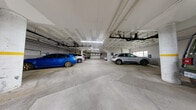This single-level enclosed parking garage provides a generously sized, functional space designed to accommodate a substantial number of vehicles across clearly marked stalls delineated by yellow painted lines on a concrete floor. The structure features a coffered concrete ceiling finished in white, with an extensive network of exposed pipes, conduits, and mechanical systems running overhead — all painted uniformly to maintain a cohesive appearance. Fluorescent strip lighting is mounted throughout, ensuring consistent illumination across the entire floor plate.

Structural support columns, painted white with yellow safety banding, are positioned at regular intervals and help define traffic flow lanes within the garage. Access is facilitated by at least one overhead roll-up garage door, and interior pedestrian access points with secure doors connect to the building beyond. A stairwell entry is also visible, reinforcing connectivity to the broader building.

Along certain perimeter walls, small windows allow modest amounts of natural light to filter in, supplementing the artificial lighting. Informational and regulatory signage is posted at multiple locations throughout the space. A fire extinguisher and floor drains are present, reflecting standard safety and drainage provisions. The overall environment is utilitarian and well-organized, suited for secure, covered vehicle storage.