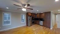 This single-floor space combines functional design with warm, inviting aesthetics. The open-concept layout seamlessly connects the living area to the kitchen, creating a cohesive and practical environment. Hardwood flooring in a rich honey tone runs throughout, lending warmth and continuity to the space.

The kitchen is a standout feature, outfitted with dark cherry wood cabinetry that extends to the upper walls, offering generous storage. A full suite of stainless steel appliances includes a French door refrigerator with an ice and water dispenser, a gas range, and a dishwasher. The dark tile backsplash provides a sharp contrast against the cabinetry, while recessed lighting ensures the area is well-lit. A sink is positioned beneath upper cabinets, maximizing workspace efficiency.

Crown molding adds a classic architectural touch along the ceiling perimeter, complementing the recessed lighting and a centrally mounted ceiling fan with a light fixture that serves the open living area. A window with horizontal blinds allows natural light to filter in, brightening the space. The overall ambiance is clean, modern, and well-appointed.