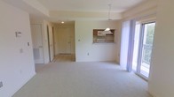 This single-floor space features an open and airy layout designed for comfortable everyday living. The main living area is finished with wall-to-wall carpeting in a neutral tone, creating a clean and cohesive feel throughout. A large window or sliding glass door along one wall allows natural light to fill the space, contributing to a bright and welcoming ambiance.

A defining feature of the layout is the pass-through kitchen opening, which connects the kitchen to the living area. The kitchen is equipped with wood-toned cabinetry, an under-cabinet range hood, and a countertop that extends to form a convenient bar or serving ledge. This open connection between the kitchen and living space enhances the sense of flow and functionality.

Beyond the main living area, a tiled entryway or hallway leads toward additional rooms, including what appears to be a closet and at least one additional door, suggesting bedroom or utility access. The transition from tile to carpet clearly delineates the entry zone from the main living space. Neutral wall colors and simple, practical finishes give the space a versatile, move-in-ready character.