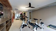 The Spin Room is a purpose-built fitness space designed to support a variety of cardio and strength training activities. The room is anchored by a row of professional-grade Spinning bikes arranged to accommodate group cycling sessions, providing ample space between each bike for comfortable use. Recessed lighting and a sleek ceiling fan overhead ensure the environment remains well-lit and properly ventilated during intense workouts. The neutral-toned walls and light flooring create a clean, modern aesthetic that contributes to an energizing atmosphere. Along one wall, a fully stocked vertical rack holds an assortment of medicine balls in varying weights, offering versatile options for functional strength training. Step platforms are also available, adding further variety to workout routines. A door leads to an adjoining area, suggesting convenient access to additional amenities or facilities. The overall layout is thoughtfully organized to maximize usability while maintaining an open, uncluttered feel, making the space suitable for both individual training sessions and guided group fitness classes.