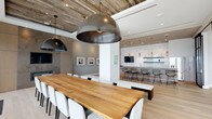 This single-floor space showcases a thoughtfully curated contemporary design that seamlessly integrates contrasting materials — raw reclaimed wood, polished marble, and clean white finishes — to create a warm yet refined aesthetic throughout.

The open-plan layout connects the living, dining, and kitchen areas cohesively. The dining area features a substantial live-edge wood table paired with white upholstered chairs, anchored beneath striking oversized dome pendant lights. A reclaimed wood-planked ceiling adds organic texture overhead, complementing the light hardwood flooring underfoot.

The kitchen is a standout element, featuring a dramatic floor-to-ceiling marble slab backsplash with bold veining that serves as a focal point. Sleek, handle-free cabinetry in warm neutral tones lines the walls, while a mixed-material island — combining concrete and reclaimed wood — provides bar seating. Cylindrical gold pendant lights add a refined finishing touch.

The living area is anchored by a striking feature wall composed of alternating reclaimed wood planks and mirrored strips housing an integrated television. Plush cream sofas and armchairs are arranged for comfortable seating, while expansive floor-to-ceiling windows flood the space with natural light and offer broad panoramic views of the surrounding cityscape.