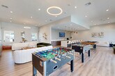 Located in Houston, TX, this expansive single-floor community amenity space offers a thoughtfully designed environment built for both socializing and wellness. The welcoming lobby greets visitors with soaring ceilings, large glazed entry doors, light hardwood flooring, and a striking marble-topped reception counter paired with warm wood cabinetry. Curated artwork and track lighting complete the polished first impression. Adjacent to the entrance, a refreshment nook features a geometric gold accent wall, a beverage station, built-in shelving, and a beverage cooler. The open-concept clubroom is the heart of the space, anchored by plush, curved seating in cream and forest green, a leather ottoman, and a large flat-screen TV. Overhead, a circular halo pendant light adds a contemporary focal point. This social hub flows into a game zone with a full-size billiards table and foosball tables, alongside a long marble-topped bar counter illuminated by gold pendant lights — ideal for gatherings. The fitness center is divided into two well-lit areas: one housing cardio equipment including treadmills, ellipticals, and a cable machine, and the other a strength-training room with a squat rack, free weights, and ceiling fans. A dedicated package locker room provides a secure, convenient parcel solution. Throughout, light hardwood floors, recessed lighting, and a neutral palette create a cohesive, modern ambiance.