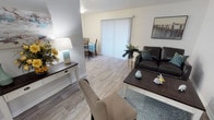 This single-floor residence offers a thoughtfully organized layout with clearly defined living spaces throughout. The living room features light wood-look flooring, neutral walls, and a large sliding glass door dressed with vertical blinds, allowing natural light to fill the room. A framed landscape painting serves as a focal point on the main wall, complementing the space's calm, coastal-inspired palette.

Adjacent to the living area, the dining space accommodates a round table with seating for four and benefits from direct access to the sliding door, enhancing the sense of openness. The kitchen is efficiently designed with white cabinetry, granite-style countertops, wood-look flooring, and a full suite of white appliances including a refrigerator, electric range, microwave, and double sink.

Both bedrooms feature plush carpeting, ceiling fans with light fixtures, and windows with blinds that provide natural light and privacy. One bedroom includes additional dresser storage and a closet. The full bathroom presents a clean, white aesthetic with a shower-tub combination, white cabinetry with overhead storage, a granite-style vanity, and wood-look flooring, completing the unit's consistent, well-maintained interior.