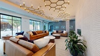 Located at 534–550 Benton Ave in Nashville, TN, this single-floor amenity lounge is a thoughtfully designed communal space that balances sophisticated aesthetics with comfortable functionality. The centerpiece of the space is a striking series of gold hexagonal chandelier installations suspended from the elevated ceilings, casting warm, ambient light throughout the open-concept interior.

The main lounge area features multiple seating arrangements with rich cognac and taupe leather sofas, accent pillows, and round coffee tables, creating an inviting atmosphere for both socializing and relaxing. A sleek linear fireplace set into a textured accent wall adds warmth and visual interest to the space.

Adjacent areas include a high-top bar seating section adorned with framed artwork, ideal for casual work or social gatherings, and a foosball table that adds a recreational touch to the layout. A dedicated seating alcove with deep navy accent chairs and floor-to-ceiling black-framed windows offers sweeping views of Geodis Park stadium directly across the street — a distinctive and dynamic vantage point.

The flooring transitions between patterned carpet with gold linear accents and wide-plank luxury vinyl tile, lending texture and visual flow throughout. Lush indoor plants are strategically placed to soften the contemporary design. An elevator vestibule and glass-partitioned corridor complete the well-appointed layout.