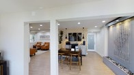 This single-floor space offers an open, thoughtfully designed layout that seamlessly connects its functional areas with a cohesive and contemporary aesthetic. The main area features smooth light wood flooring that runs throughout, creating a warm and unified feel across the space.

A central workspace anchors the interior, highlighted by a rustic dark wood table paired with matching wooden chairs, positioned in front of a distinctive geometric patterned accent wall in warm gold and white tones. A gray credenza beneath the accent wall provides practical storage, while a pair of sleek black pendant lights add a modern touch overhead. A series of abstract artwork panels adorns the accent wall, contributing to the curated, professional atmosphere.

Adjacent to the main workspace, a comfortable lounge area is defined by a pair of rich cognac leather chairs arranged on a neutral area rug, with natural light filtering in from a nearby window. Decorative bamboo plants add a natural, organic element to the space.

Recessed lighting throughout ensures even, well-distributed illumination across all areas. A frosted glass entry door allows diffused natural light into the space while maintaining privacy. Structural columns provide subtle architectural definition between zones without interrupting the open flow. Overall, this space strikes a balance between professional functionality and inviting, stylish design.