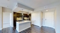 This single-floor residence offers a thoughtfully designed open-concept layout that seamlessly blends style and functionality. The kitchen serves as the heart of the space, anchored by a sleek central island with a crisp white countertop and an integrated sink, providing ample prep and gathering space. Rich, dark espresso-toned cabinetry lines the walls, offering generous storage, while a neutral tile backsplash adds visual texture and depth. Stainless steel appliances, including a refrigerator, microwave, and range, lend a contemporary, professional-grade feel to the culinary space. Pendant lighting hangs above the island, casting warm, focused light that complements the recessed ceiling fixtures throughout. The flooring features wide-plank hardwood-style planks in a warm gray tone, flowing continuously through the kitchen and into the adjoining areas, creating a cohesive and grounded aesthetic. A set of double shutter-panel doors provides access to additional storage, while a secondary door leads to an entryway or adjacent room. Neutral gray walls and clean white trim maintain a bright, airy atmosphere throughout, making this a well-appointed and visually balanced living space.