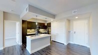 This single-floor residence offers a thoughtfully designed open-concept layout that seamlessly blends style and functionality. The kitchen serves as the heart of the space, anchored by a sleek central island with a crisp white countertop and an integrated sink, providing ample prep and gathering space. Rich, dark espresso-toned cabinetry lines the walls, offering generous storage, while a neutral tile backsplash adds visual texture and depth. Stainless steel appliances, including a refrigerator, microwave, and range, lend a contemporary, professional-grade feel to the culinary space. Pendant lighting hangs above the island, casting warm, focused light that complements the recessed ceiling fixtures throughout. The flooring features wide-plank hardwood-style planks in a warm gray tone, flowing continuously through the kitchen and into the adjoining areas, creating a cohesive and grounded aesthetic. A set of double shutter-panel doors provides access to additional storage, while a secondary door leads to an entryway or adjacent room. Neutral gray walls and clean white trim maintain a bright, airy atmosphere throughout, making this a well-appointed and visually balanced living space.