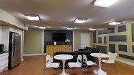 This single-floor space functions as a comfortable lounge or common area, thoughtfully arranged to accommodate both relaxed socializing and casual gatherings. The room features warm hardwood flooring that runs throughout, providing a welcoming foundation that contrasts nicely with the cooler tones of the walls.

The focal point of the main wall is a mounted flat-screen television, set against a dark accent panel that adds visual depth and a contemporary touch. Beneath the TV sits a natural wood dresser-style console, styled with a floral arrangement and decorative accents, lending a homey character to the space.

Seating is thoughtfully varied: a pair of sleek black leather sofas offer ample lounging space along the perimeter, while several round white bistro-style tables paired with modern white chairs are arranged throughout the open floor area, creating a relaxed café-like atmosphere suited for conversation or casual work.

Overhead, recessed fluorescent light panels provide broad, even illumination across the room. The walls feature a mix of neutral tones with dark rectangular accent panels, contributing a structured yet informal aesthetic. A white interior door provides access to adjacent areas, completing the layout of this versatile, well-appointed communal space.