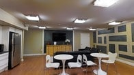 This single-floor space functions as a comfortable lounge or common area, thoughtfully arranged to accommodate both relaxed socializing and casual gatherings. The room features warm hardwood flooring that runs throughout, providing a welcoming foundation that contrasts nicely with the cooler tones of the walls.

The focal point of the main wall is a mounted flat-screen television, set against a dark accent panel that adds visual depth and a contemporary touch. Beneath the TV sits a natural wood dresser-style console, styled with a floral arrangement and decorative accents, lending a homey character to the space.

Seating is thoughtfully varied: a pair of sleek black leather sofas offer ample lounging space along the perimeter, while several round white bistro-style tables paired with modern white chairs are arranged throughout the open floor area, creating a relaxed café-like atmosphere suited for conversation or casual work.

Overhead, recessed fluorescent light panels provide broad, even illumination across the room. The walls feature a mix of neutral tones with dark rectangular accent panels, contributing a structured yet informal aesthetic. A white interior door provides access to adjacent areas, completing the layout of this versatile, well-appointed communal space.