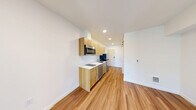 This thoughtfully designed one-bedroom, one-bathroom unit in Seattle, WA offers a smart, efficient layout that maximizes every square foot. Warm hardwood-style flooring runs continuously throughout the space, complementing bright white walls and recessed lighting for a clean, modern aesthetic.

The combined kitchen and living area forms the heart of the unit. The galley-style kitchen is outfitted with light wood cabinetry, white countertops, stainless steel appliances including a built-in microwave, dishwasher, and refrigerator, and a single-basin sink — delivering a fully functional cooking space in a compact footprint. The adjacent living area benefits from natural light streaming in through French doors that open onto a private balcony, creating an airy, open feel.

The bedroom is accessed via a sleek sliding glass-panel door, offering a sense of separation while maintaining an open and light-filled atmosphere. The bathroom features matching light wood cabinetry with a white countertop vanity, a full tub and shower, chrome fixtures, and open wall shelving. The consistent use of warm wood tones and white finishes throughout the unit creates a cohesive, contemporary look ideal for efficient urban living.