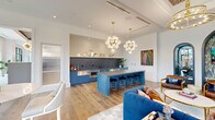 Located at Bennet at Bull Street in Columbia, SC, this thoughtfully designed single-floor space offers a generously proportioned open-plan layout that seamlessly integrates a fully equipped kitchen, multiple lounge areas, and distinct architectural features throughout.

The kitchen serves as a striking focal point, showcasing bold blue cabinetry paired with a large center island topped with a grey quartz surface and lined with black velvet bar stools. A dramatic dark marble-veined backsplash contrasts with shiplap-paneled upper walls, while globe chandelier pendants with brass fixtures illuminate the space from above. Stainless steel appliances complete the functional yet stylish setup.

The lounge areas are defined by curated furnishings, including deep tufted navy sofas, leather armchairs, and layered area rugs. A coffered ceiling with an ornate lattice detail and tiered chandelier anchors one seating zone, while floor-to-ceiling steel-framed windows flood the space with natural light and offer views to the exterior.

Distinctive arched niches adorned with botanical wallpaper and decorative lighting add an artistic, boutique-hotel quality to the space. Wide-plank hardwood flooring runs throughout, tying each zone together cohesively and contributing to the space's refined, upscale ambiance.